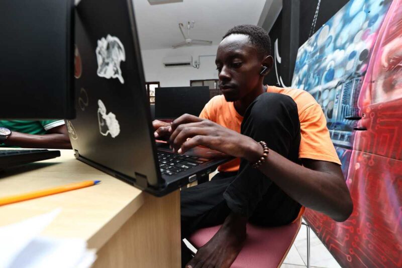 Man in orange shirt types intently on a laptop at a computer lab station.