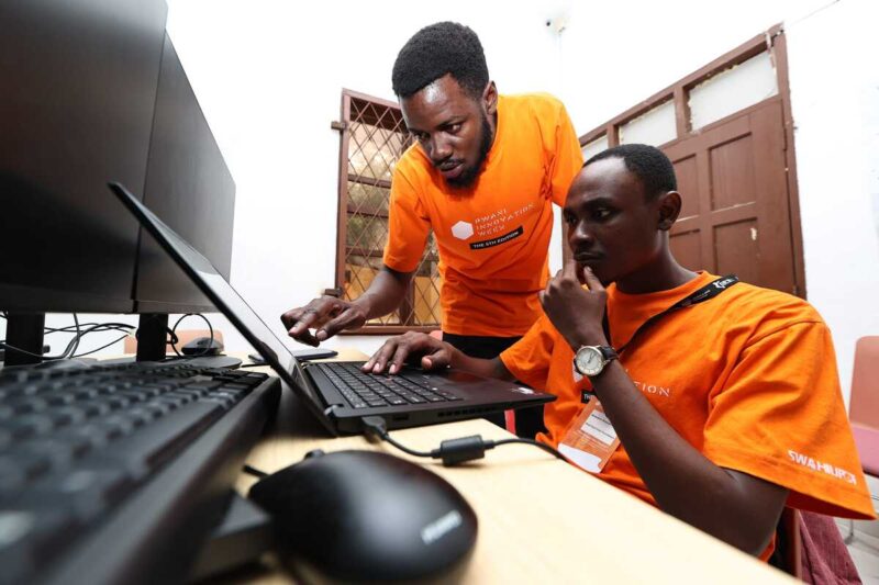 Two young men in orange shirts focus on coding together at dual-screen workstations.