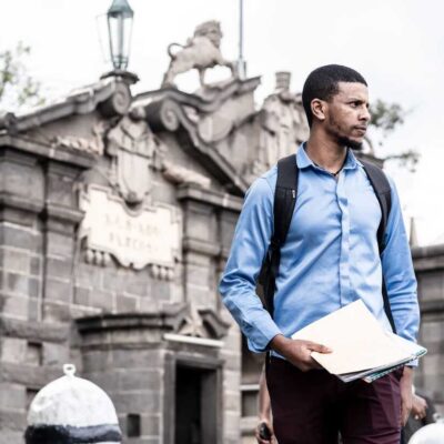 Man in blue shirt walks outside a historic stone building holding documents.