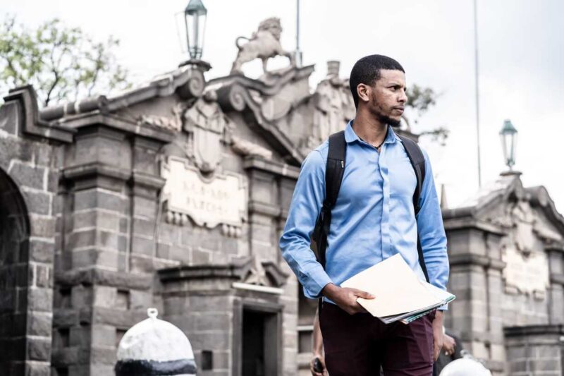Man in blue shirt walks outside a historic stone building holding documents.