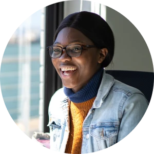 Smiling woman in glasses and a denim jacket sits by a sunny window during a conversation.