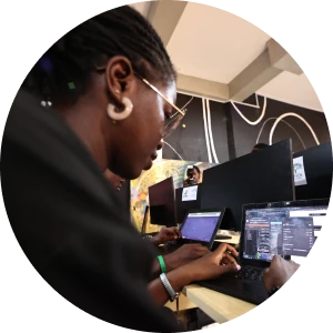 Woman wearing glasses and earrings types on a laptop in a shared workspace with multiple monitors and people coding.