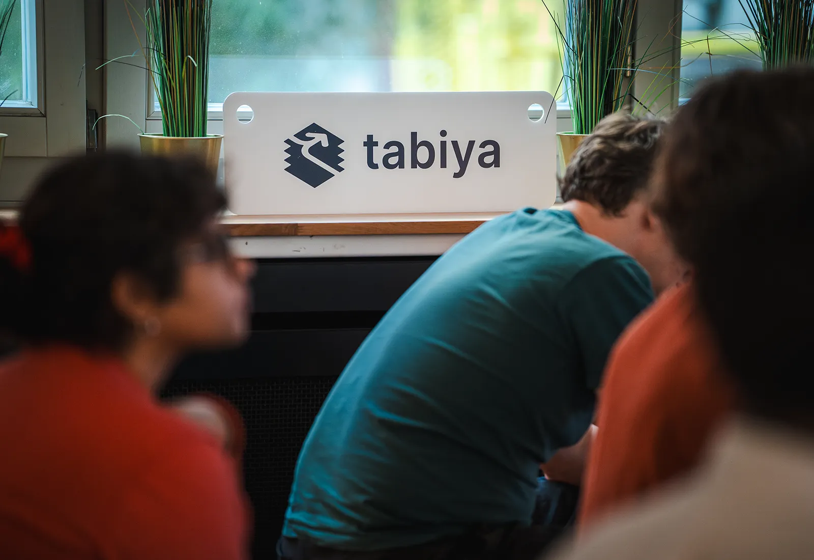 Tabiya-branded desk sign with people listening to a presentation nearby, framed by potted plants.