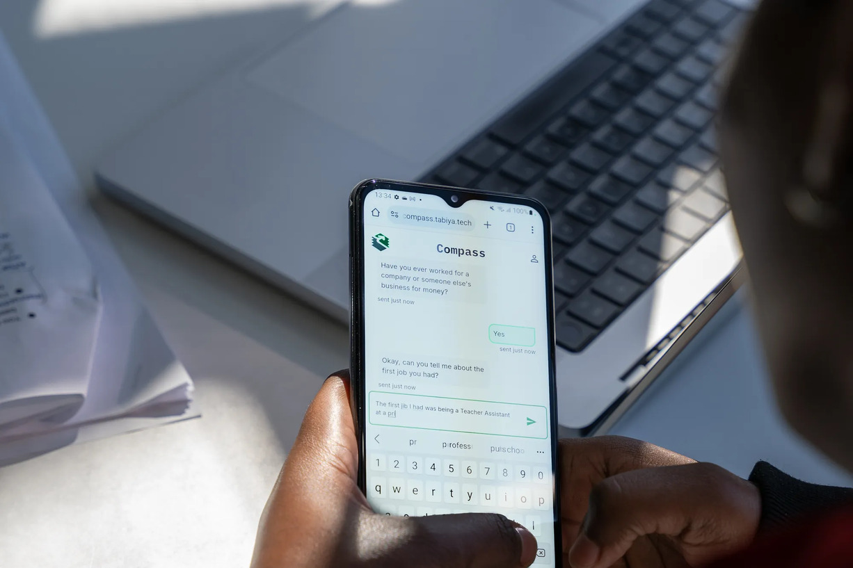 Woman using Compass chatbot app on a phone while working at a laptop.